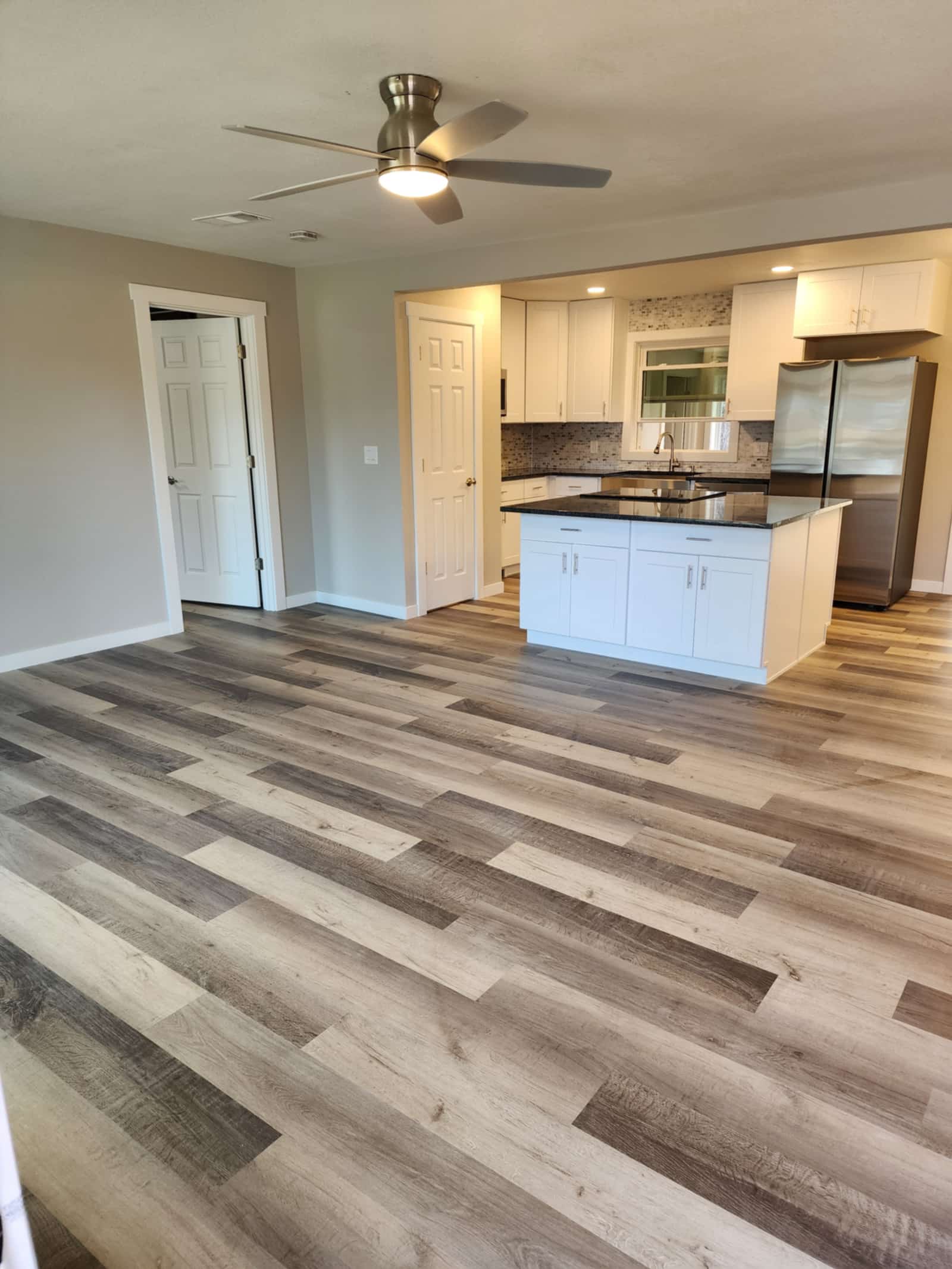 Finished kitchen with white shaker cabinets, dark granite countertops, a mosaic tile backsplash and a central island, opening onto a living space with new plank flooring